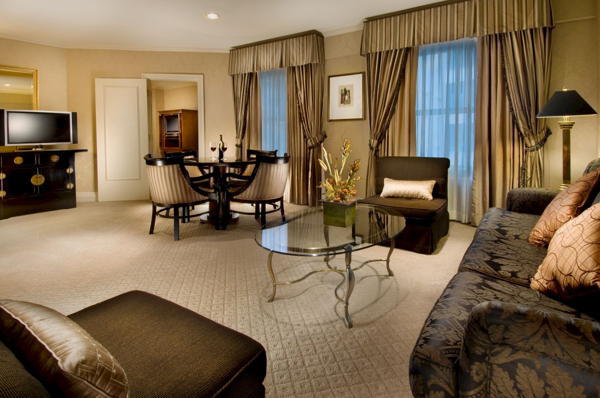 A luxurious hotel suite with a cream carpet, dark upholstered sofas, a glass coffee table, a round dining table, curtains, and soft lighting.