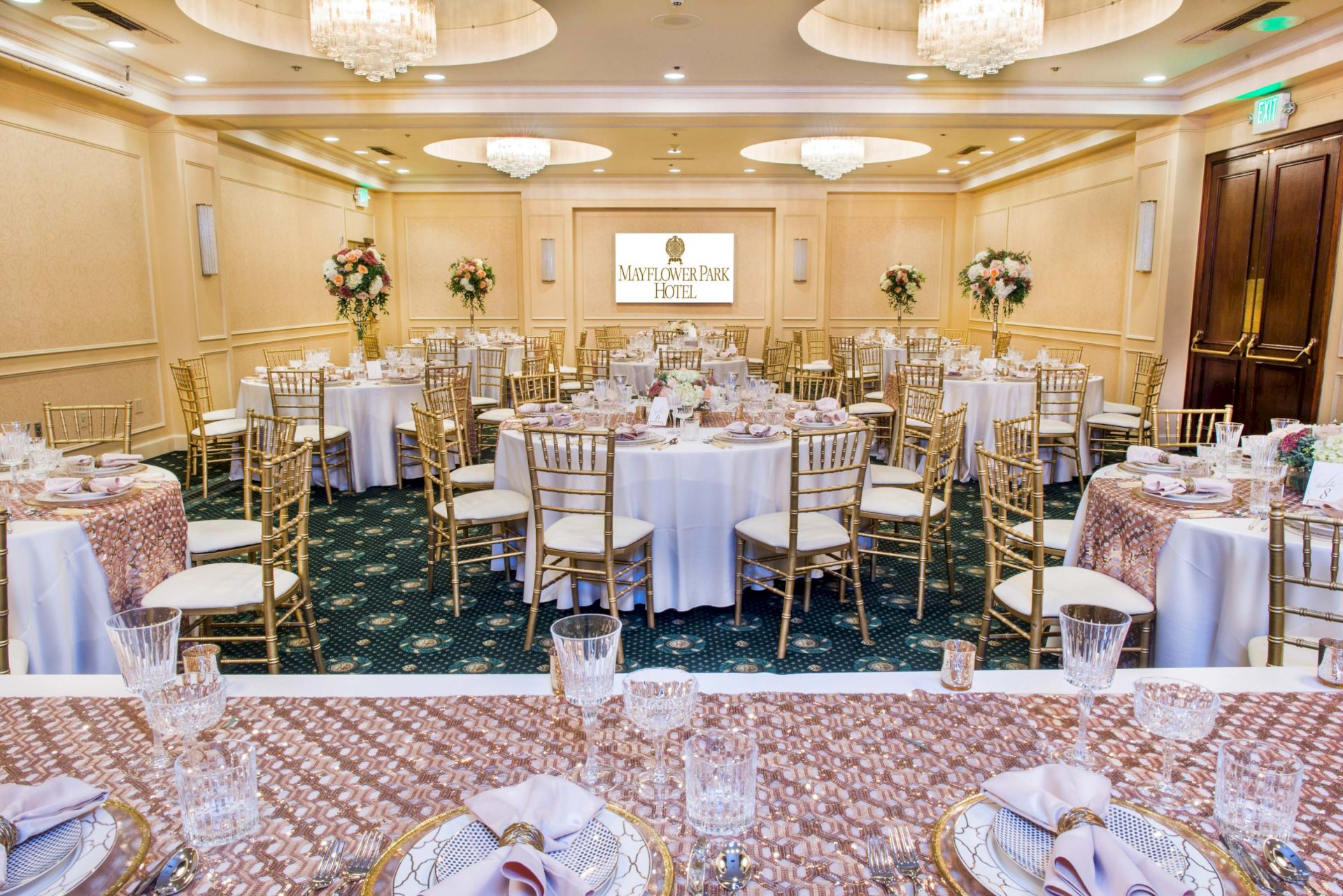 Elegant banquet hall with round tables, gold chairs, crystal glassware, floral centerpieces, and a Mayflower Park Hotel sign.