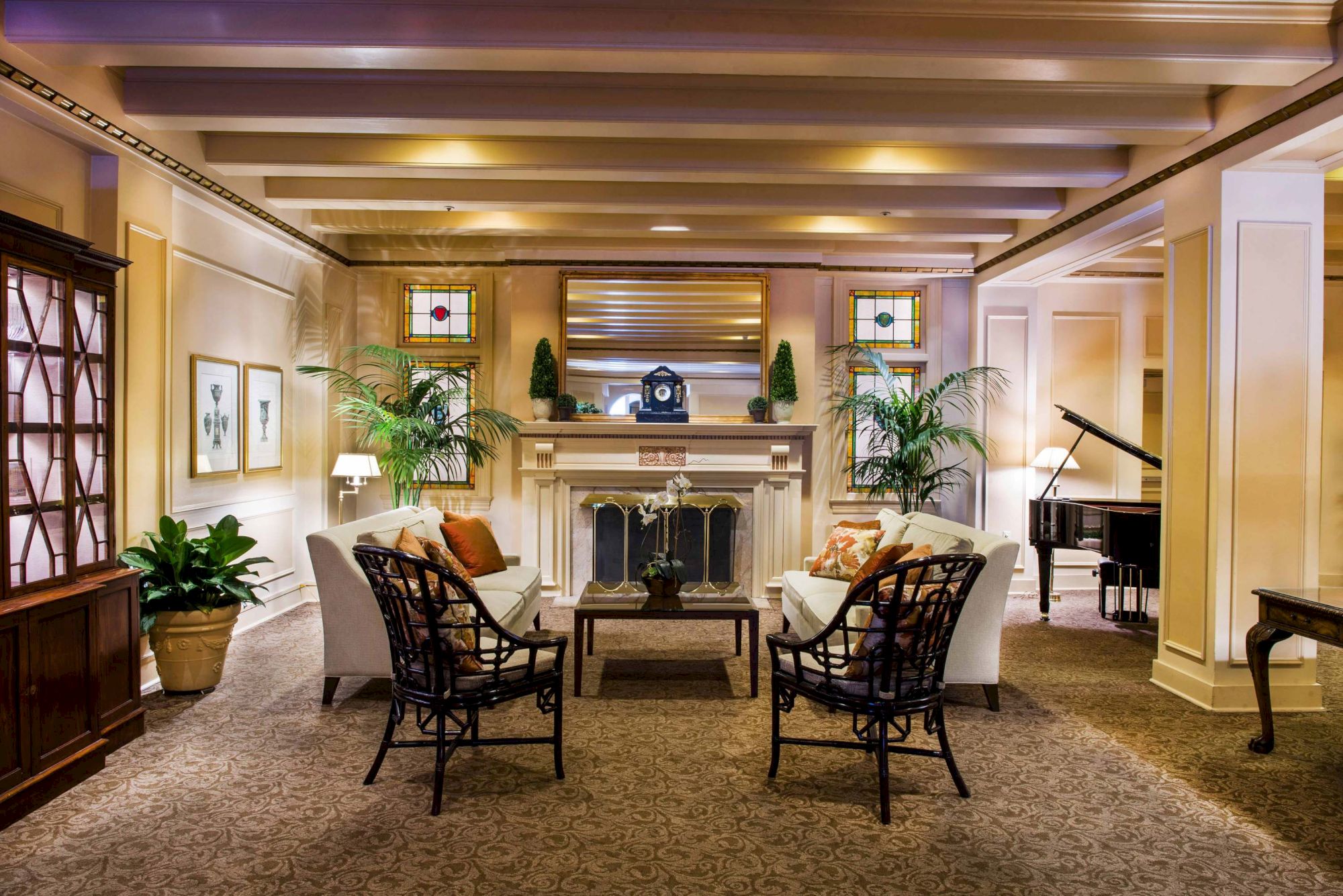 A cozy hotel lobby with a stone fireplace, two white sofas facing each other, wicker chairs, potted plants, warm lighting, and a grand piano in the corner.