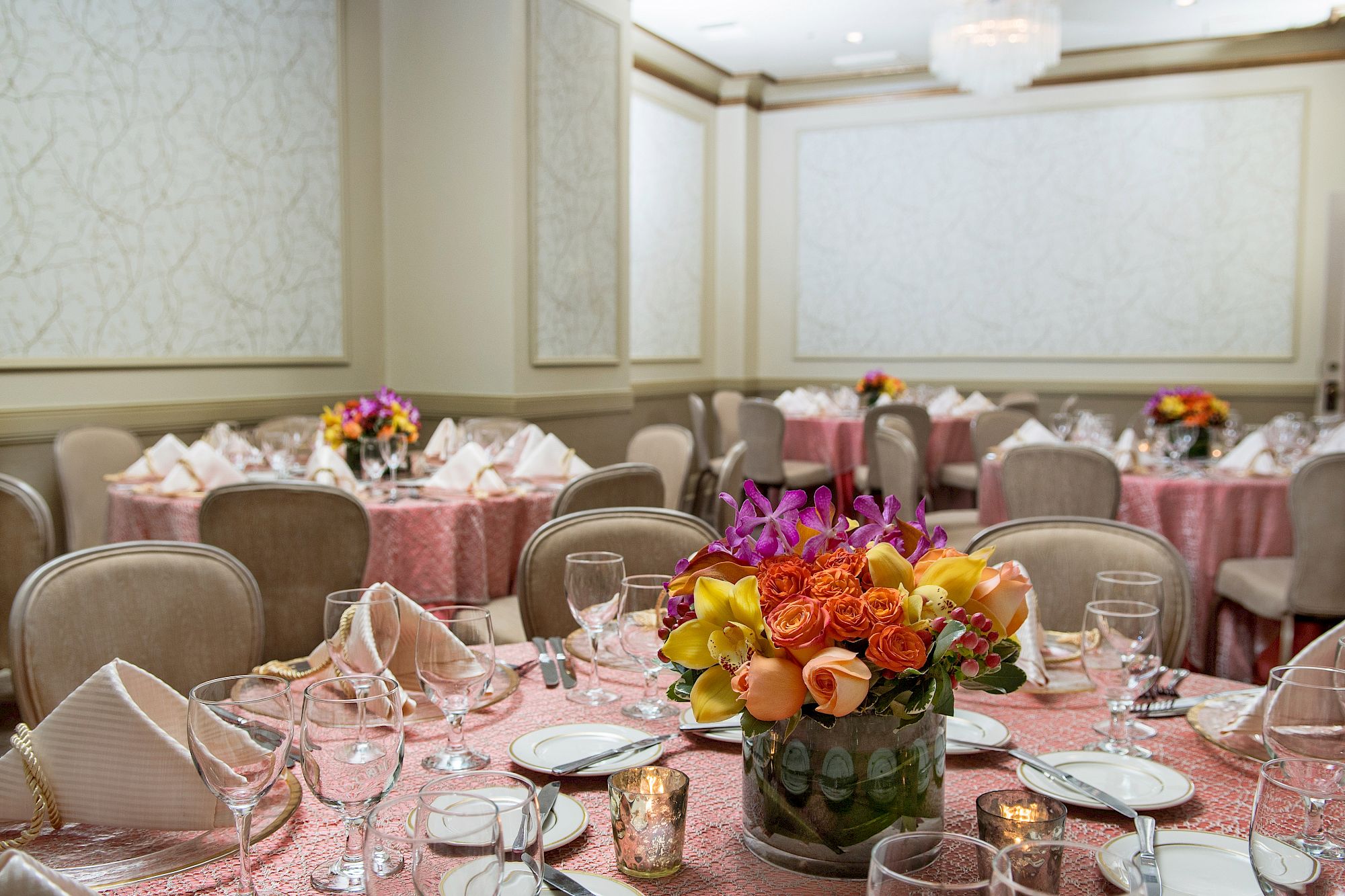 A banquet hall with round tables dressed in pink tablecloths, flower centerpieces, and neatly arranged place settings for an event.