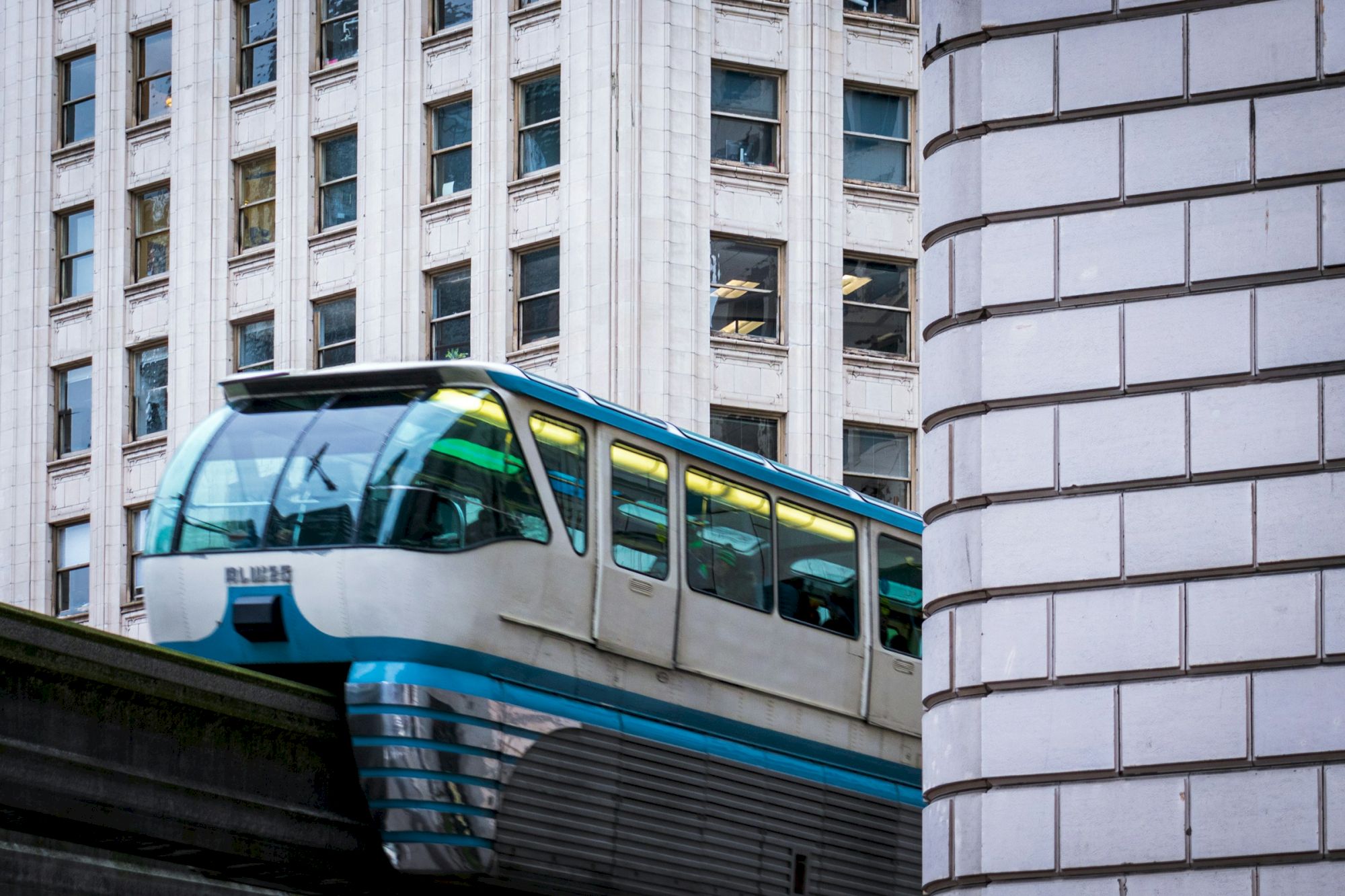 A monorail or elevated train peeks between buildings, with a blue stripe and curved windows, emerging between white-brick facades.
