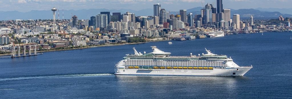 A cruise ship sails past a bustling city skyline with tall buildings, a calm harbor, and distant mountains under a clear blue sky.