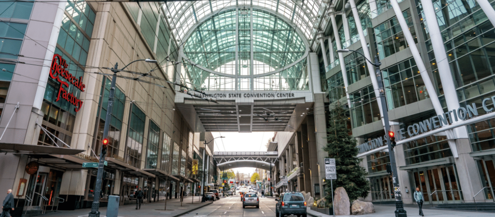 Seattle-style city street scene at a convention center, glass atrium, tall buildings, cars, pedestrians, and a skybridge overhead.