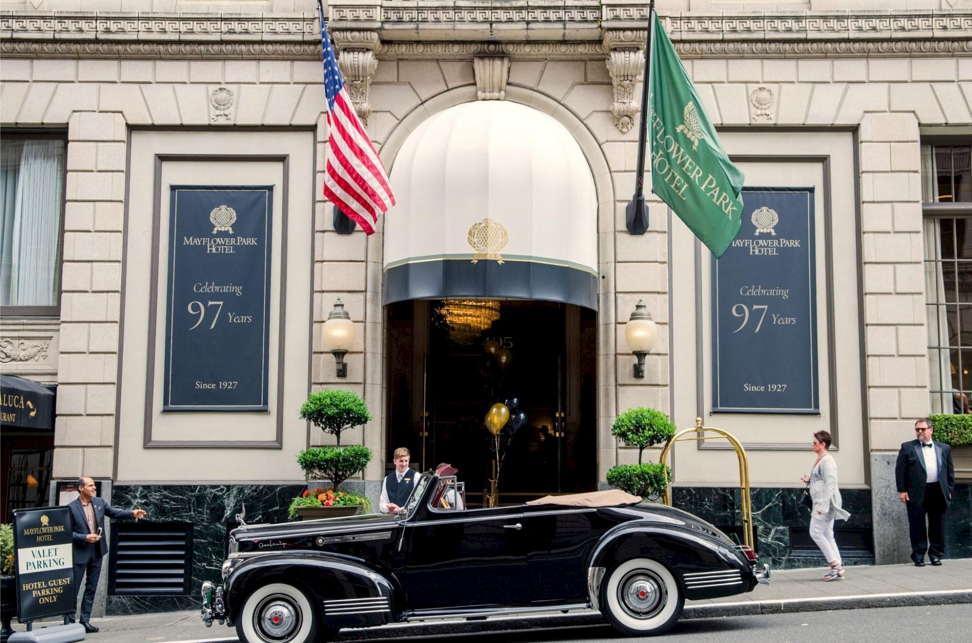 A black vintage car sits in front of a grand hotel entrance with banners and people nearby, under an American flag.