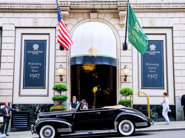 A vintage black car pulls up outside a grand hotel entrance, with flags, uniformed staff, and people strolling by on a sunny day.