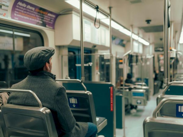 A man wearing a gray coat and hat sits alone on a mostly empty subway car, holding onto a pole and looking ahead.