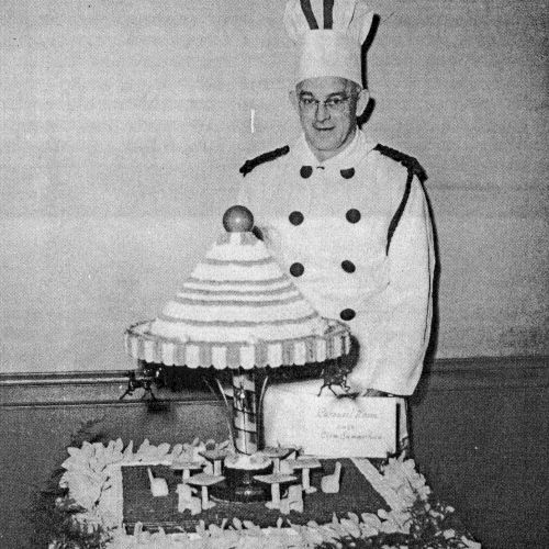 A vintage black-and-white photo of a person in a formal uniform standing beside a pie-topped, multi-layered cake on a table, labeled "Birthday."