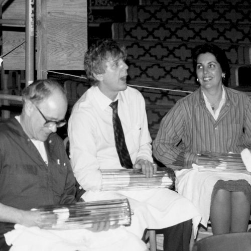 A black-and-white photo shows three men at a table with papers; one in the middle points, another writes, and a third leans in, all smiling.