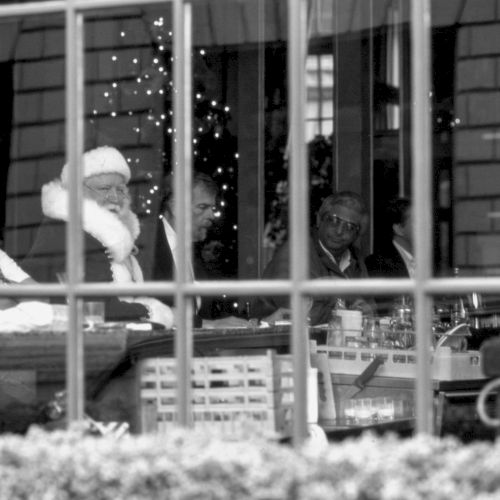 A man sits in a jail cell, leaning on a bench behind bars, with a small table visible and a windowed wall in the background.