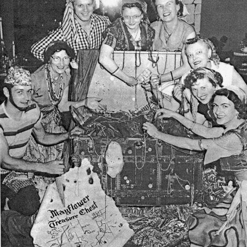 A vintage black-and-white scene of people gathered around a table, sharing food and conversation, with playful, candid poses.