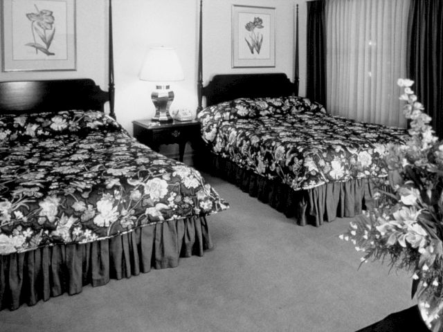 A black and white photo of a hotel room with two beds, floral bedding, and framed artwork on the walls.