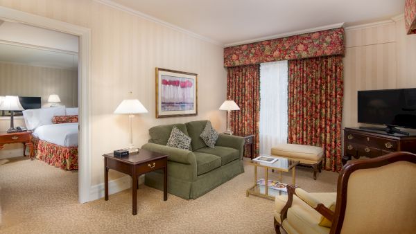 A living room with a green sofa, side tables, lamps, a coffee table, a wooden TV stand, and a floral curtain, adjoining a bedroom visible through an open doorway.
