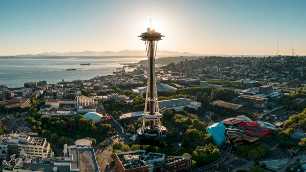 Seattle&rsquo;s Space Needle rise above a coastal cityscape at sunset, with water, hills, and urban buildings stretching toward the horizon.