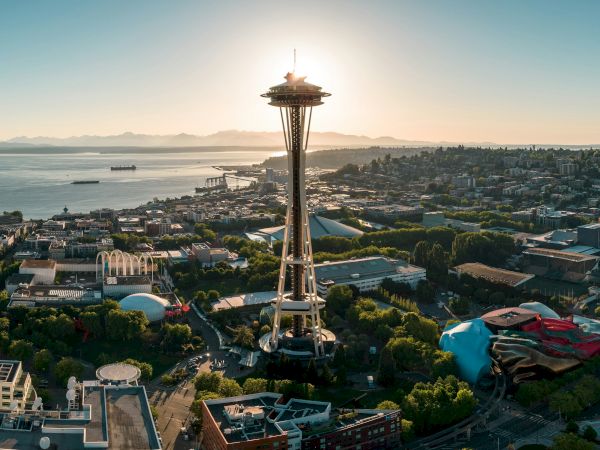 Seattle&rsquo;s Space Needle rise above a coastal cityscape at sunset, with water, hills, and urban buildings stretching toward the horizon.