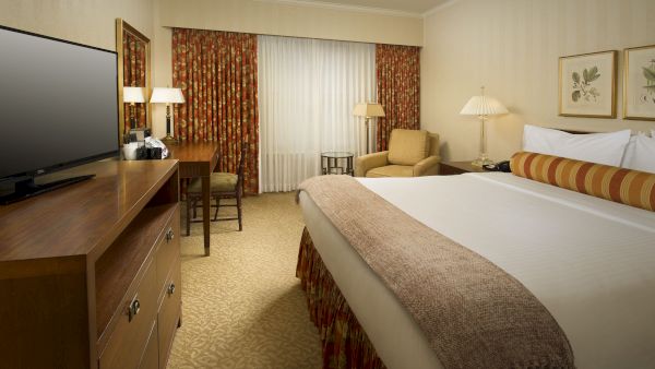 A hotel room with a large bed, beige decor, two lamps, a seating area, curtains, and a TV on a dresser, all in a warm, cozy style.