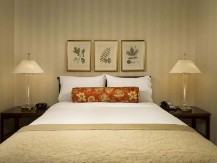 A neatly made hotel bed with white linens, beige bedspread, two lamps, and botanical wall art above the headboard, framed pictures.