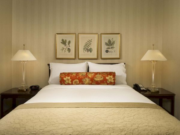 A neatly made hotel bed with white linens, beige bedspread, two lamps, and botanical wall art above the headboard, framed pictures.