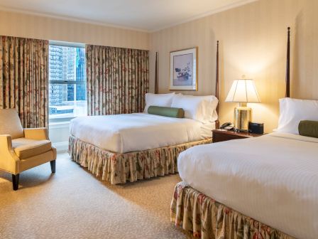 A hotel room with two queen beds, floral bed skirts, beige walls, a nightstand with a lamp, a chair by the window, and draped curtains.
