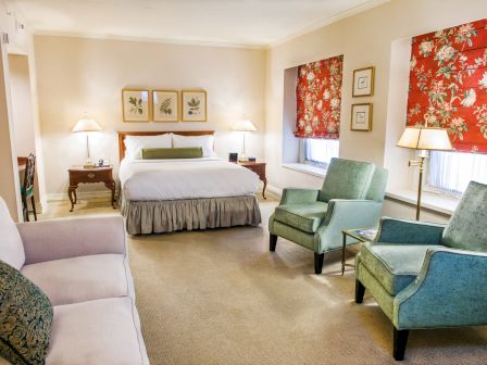 A deluxe hotel suite with a king bed, two green armchairs, a sofa, lamps, and floral red window coverings, creating a cozy, classic room.