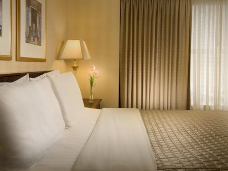 A hotel room with a neatly made bed, white linens, bedside lamp, framed art, and a window with heavy curtains letting light in, calm and tidy.