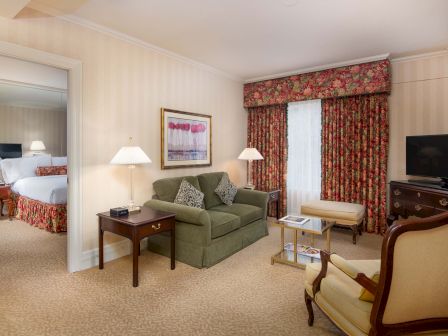 A cozy hotel suite with a green sofa, side tables, lamps, framed art, floral curtains, a desk, and a TV, separated from a separate bedroom.
