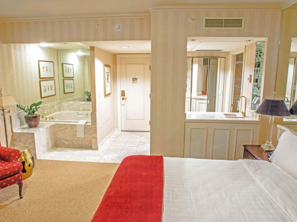 A hotel room with a bed in the foreground, red throw blanket, red chair, and a desk; bathroom area with mirrors, sink, and a hallway leading to a door.