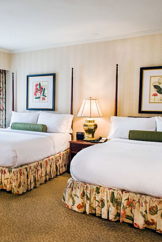 Two double beds with floral bed skirts and matching throw pillows, a seating chair with ottoman, nightstands, lamps, and framed botanical art in a beige hotel room.