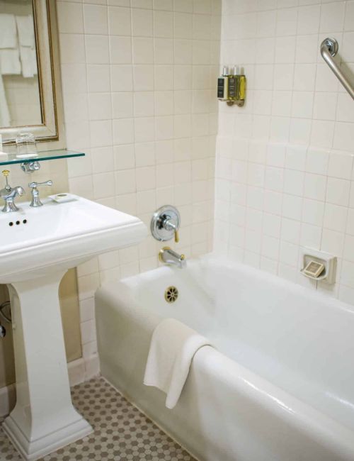 A small vintage-style bathroom with a pedestal sink, a toilet, and a bathtub with a grab bar; tile walls and a mirror above the sink, ending with a curtain.