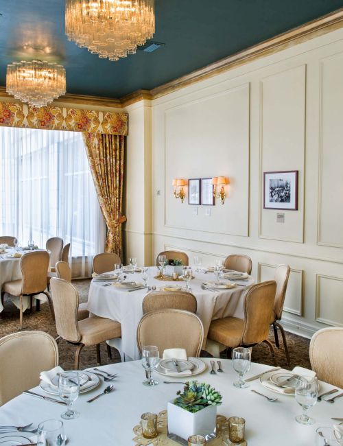 A bright, elegant dining room with round white-clothed tables, beige chairs, wall paneling, chandeliers, and large curtained windows.