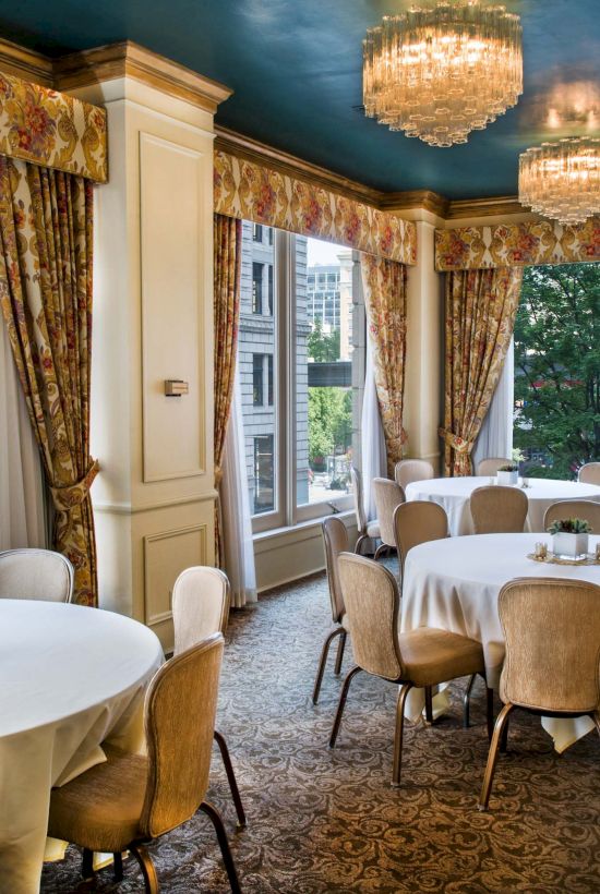 Elegant dining room with round tables, beige chairs, white tablecloths, ornate curtains, chandeliers, and large windows looking out to greenery.