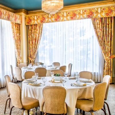 Elegant dining room with a round table set for a formal meal, cream chairs, floral curtains, crystal chandeliers, and soft natural light.