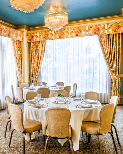 Elegant dining room with a round table set for a formal meal, cream chairs, floral curtains, crystal chandeliers, and soft natural light.