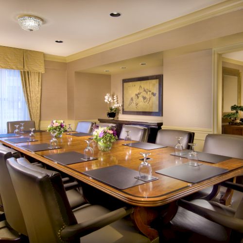 A formal boardroom with a long wooden table, leather chairs, glassware, flowers, and warm beige d&eacute;cor.
