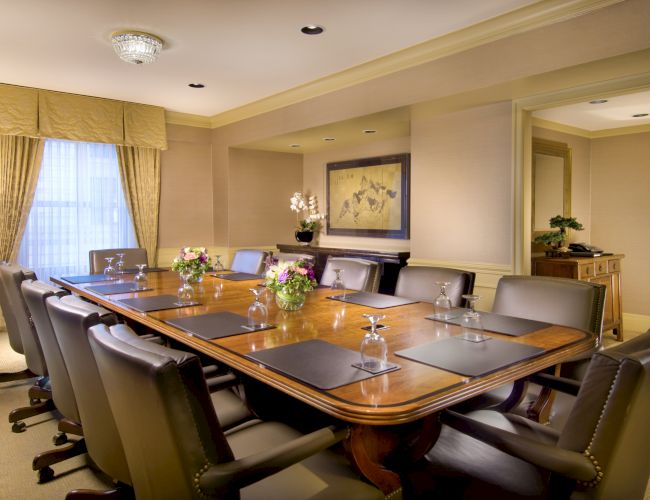 A formal boardroom with a long wooden table, leather chairs, glassware, flowers, and warm beige d&eacute;cor.