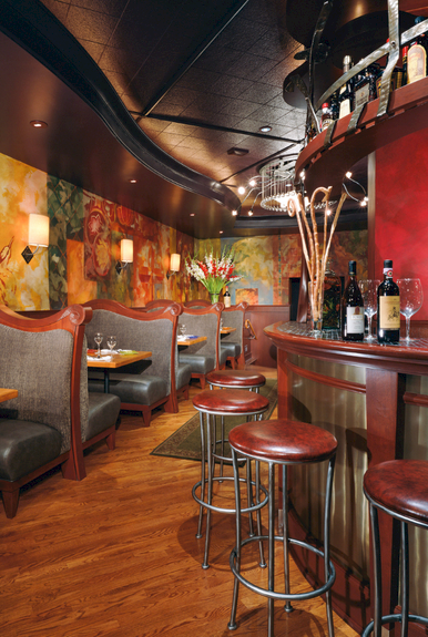 A cozy bar with a curved red counter, tall stools, warm wood flooring, and colorful mosaic wall panels behind intimate booths, bottles on display.
