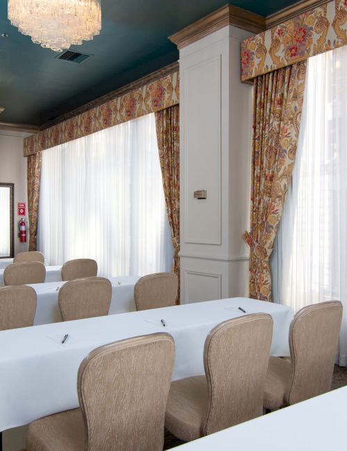 A conference room with white tablecloths, beige chairs, large windows with sheer curtains, ornate drapes, wall-mounted TV, and chandeliers.