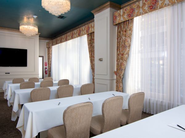 A conference room with long white-clothed tables, beige chairs, large windows with sheer curtains, and elegant chandeliers in a bright setting.