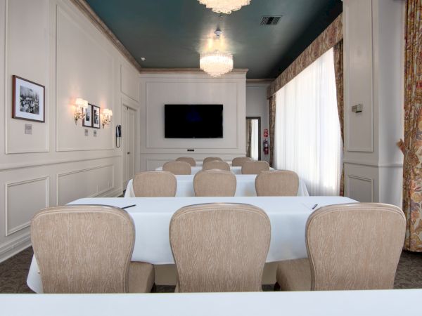 A small conference room with white paneled walls, beige chairs around white tables, a wall-mounted TV, and chandeliers, bright with curtains.