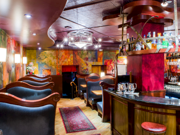 A cozy, stylish bar with curved leather booths, colorful mural walls, warm lighting, a curved counter, and shelves stocked with drinks.