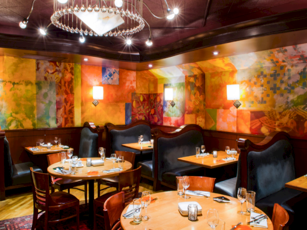 A cozy restaurant with colorful mural walls, round wooden tables, black upholstered booths, and warm lighting for a intimate dining vibe.
