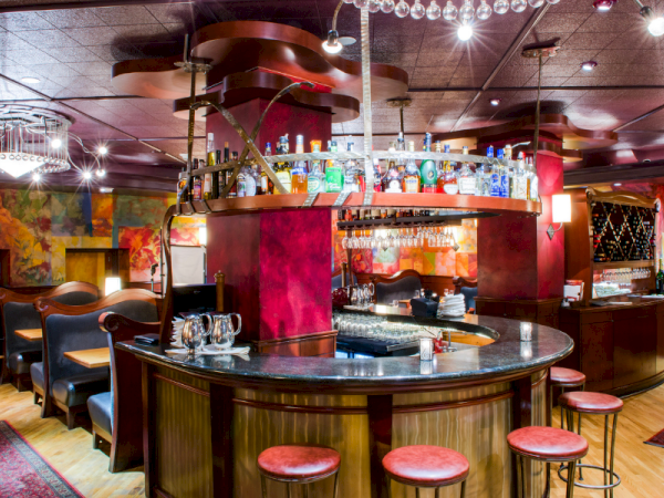 A stylish circular bar with wooden counter, red stools, glass shelves of bottles, colorful wall decor, and intimate seating seating around.