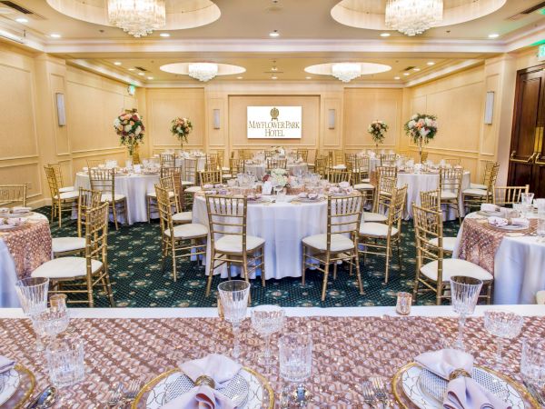 A beautifully set banquet hall with round tables, white linens, gold chairs, floral centerpieces, and a projector screen at the front for a formal event.