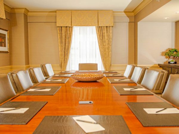A long wooden conference table with brown placemats, notepads, and a central bowl, surrounded by beige leather chairs in a formal meeting room.