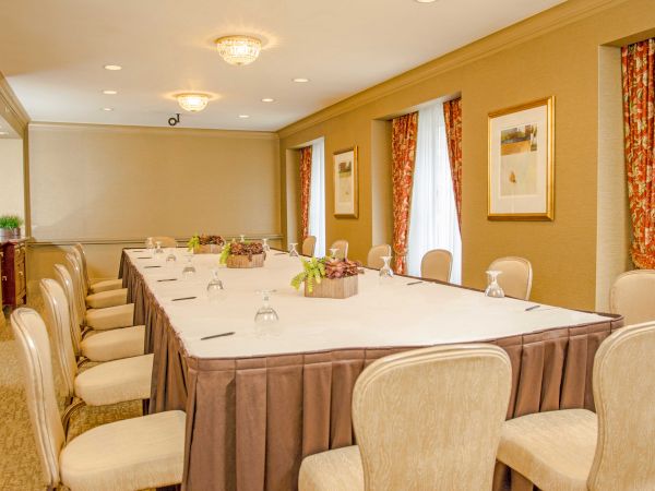 A formal conference room with a long rectangular table, beige chairs, cream walls, red drapes, framed art, soft lighting, and a neat setup.