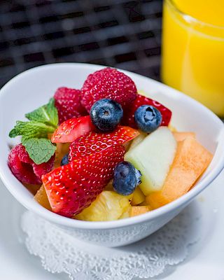 A bowl of fresh mixed fruit (strawberries, blueberries, melon, pineapple, cantaloupe) with mint, on a white plate beside orange juice.