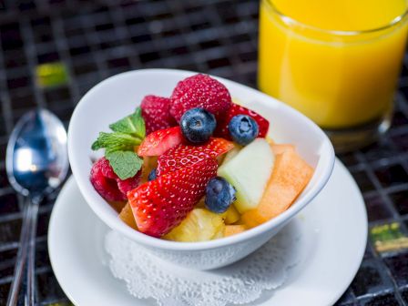 A bowl of fresh mixed fruit (strawberries, blueberries, melon, pineapple, cantaloupe) with mint, on a white plate beside orange juice.