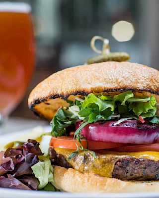 A juicy burger with greens, tomatoes, onions, and cheese on a sesame bun, plus fries and a beer in the background.