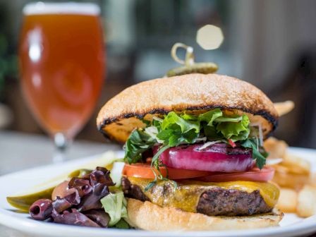 A juicy burger with greens, tomatoes, onions, and cheese on a sesame bun, plus fries and a beer in the background.