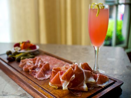 A charcuterie board with assorted cured meats on a wooden platter, plus a pink cocktail in a champagne flute beside it.
