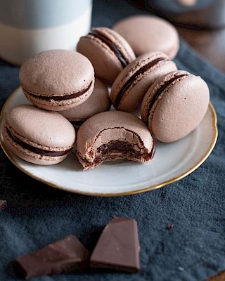 Delicate pink macarons with chocolate filling on a plate, a bite reveals rich ganache; chocolate shards and coffee cup in the background, yum.